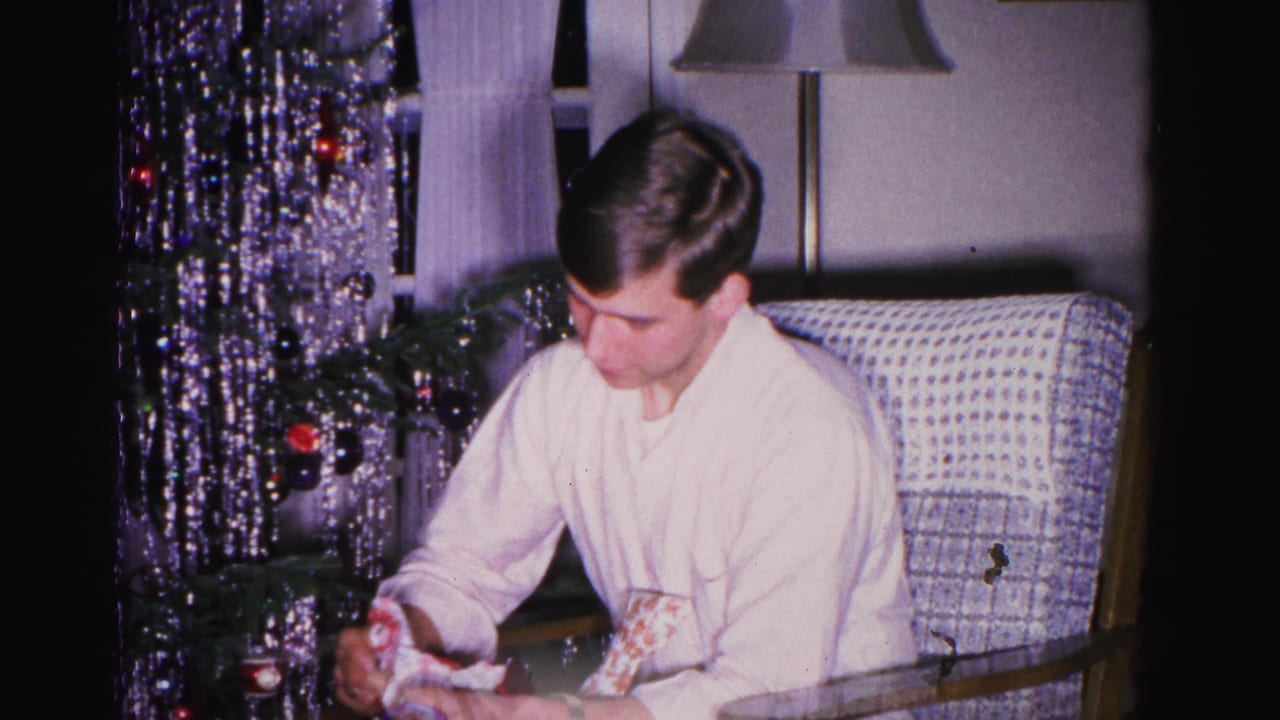 Young man opening a Christmas gift in a mid-century American living room, circa 1962, Hagerstown, Maryland; tinsel tree and beaded curtain visible at left.