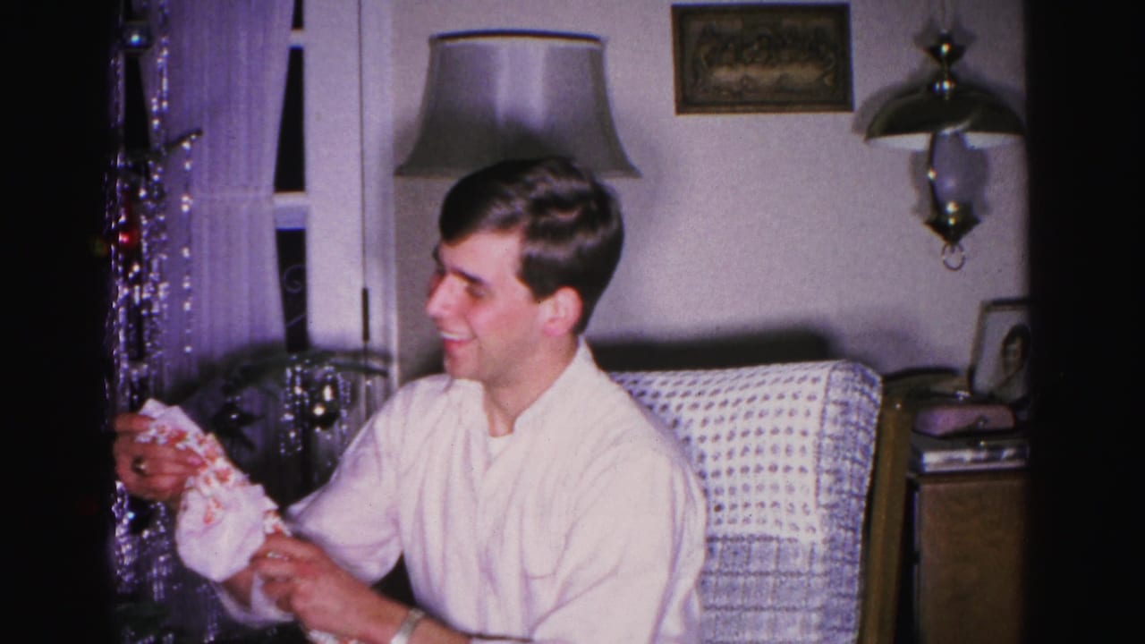 Young man smiling with unwrapped Christmas gift packaging, circa 1962; floor lamp, wall sconce, framed picture, and tinsel curtain visible in mid-century living room.