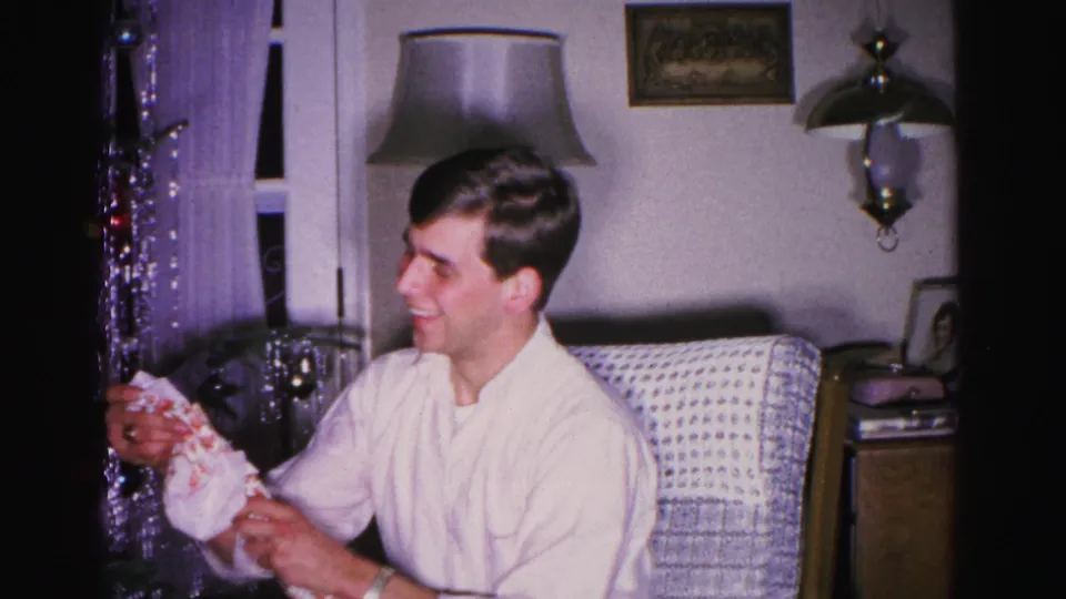Young man smiling with unwrapped Christmas gift packaging, circa 1962; floor lamp, wall sconce, framed picture, and tinsel curtain visible in mid-century living room.