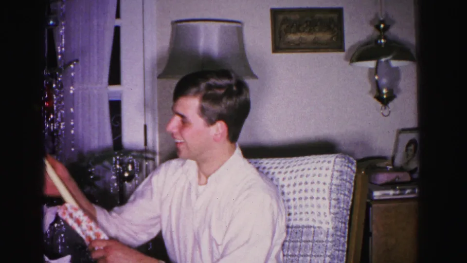 Young man in three-quarter profile holding a long-handled garden tool in a mid-century American living room, circa 1962; wood-veneer cabinet visible in background.