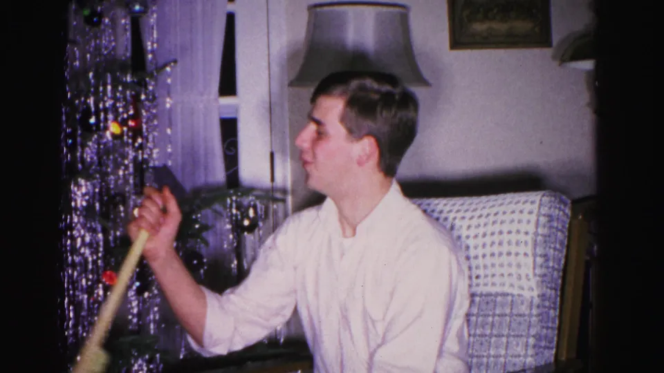 Young man smiling with a garden tool gift, circa 1962; tinsel curtain and colored Christmas tree lights visible in mid-century American home interior.