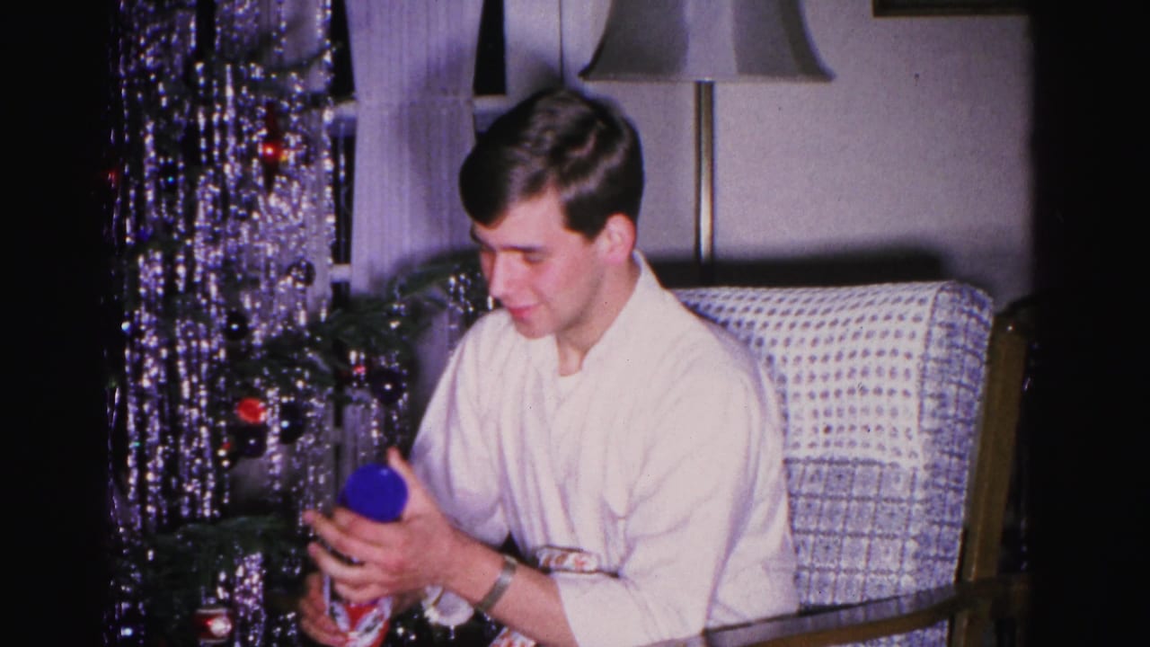 Young man in white shirt holds a blue rounded gift while seated in a woven armchair; a heavily tinsel-draped Christmas tree with red ball ornaments fills the background, ca. 1962 Hagerstown, Maryland.
