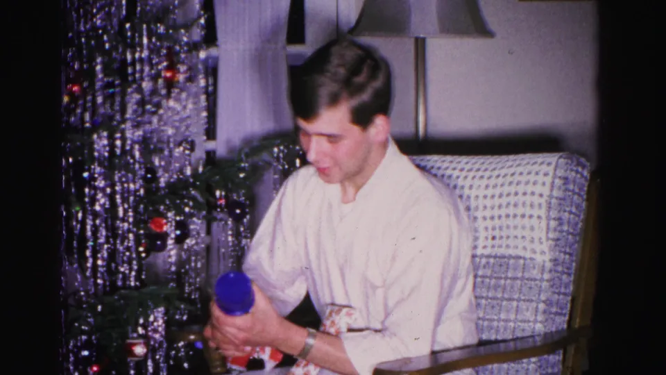 Young man leans forward to examine a blue gift, the metallic tinsel Christmas tree filling the background; living room setting, ca. 1962 Hagerstown, Maryland.