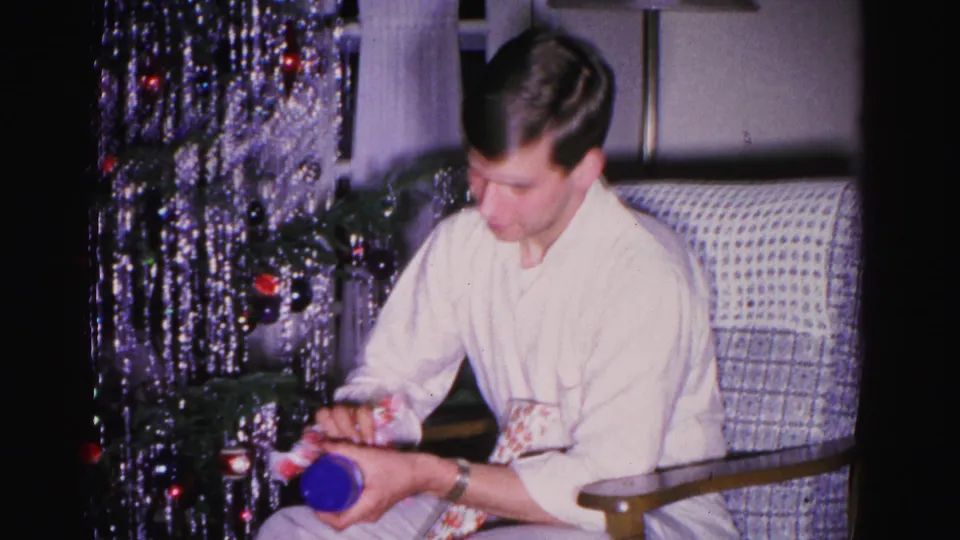 Close view of young man in white shirt holding a blue gift; silver tinsel Christmas tree and woven armchair with patterned cushion visible in background, Hagerstown, Maryland, ca. 1962.
