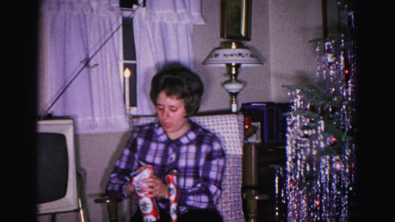 Woman in plaid shirt examines a wrapped Christmas gift; CRT TV, tinsel tree, and lamp visible in background.