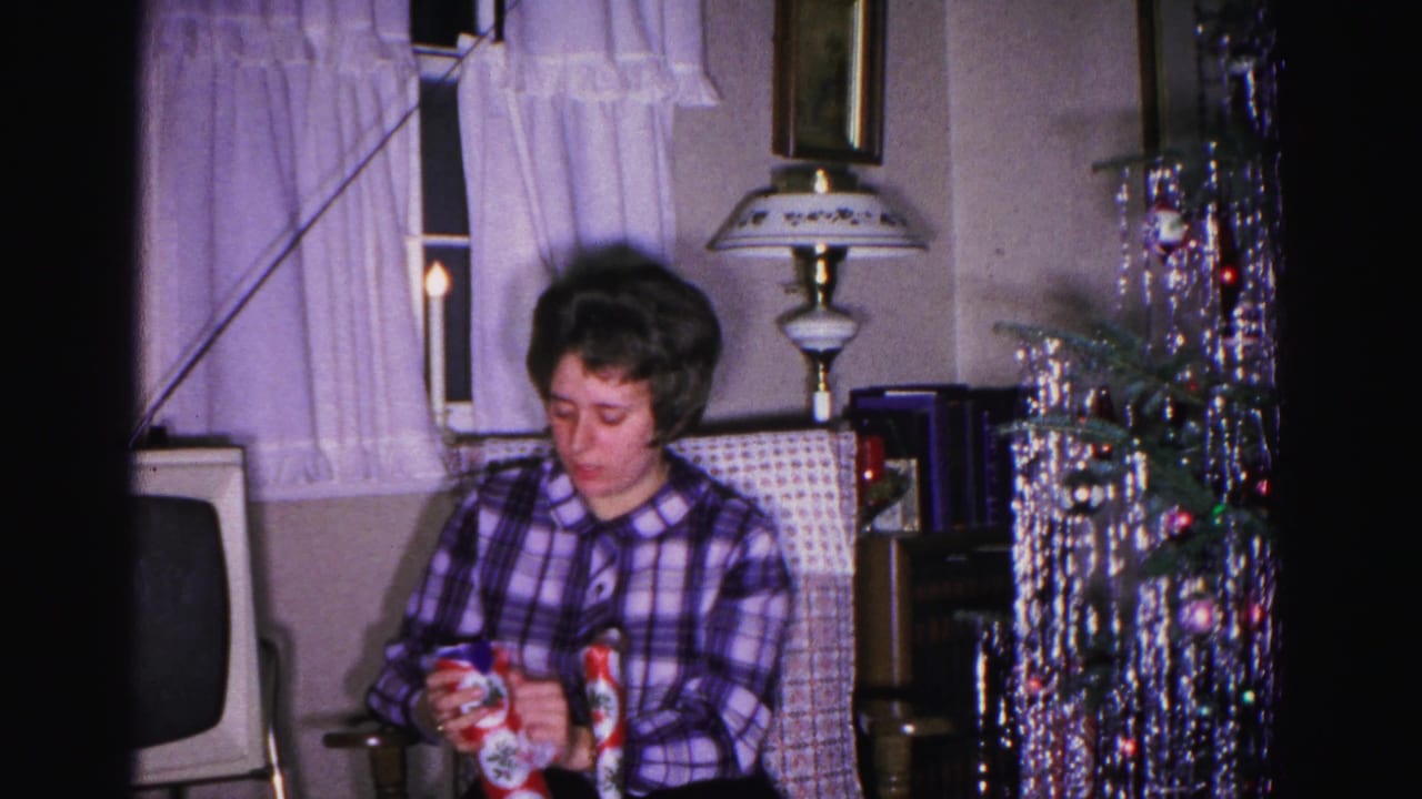 Woman examining a wrapped gift in an early 1960s living room; aluminum tinsel tree and period CRT television set prominent in background.