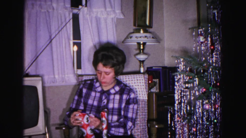 Mid-shot of a woman carefully unwrapping a Christmas gift in a 1960s American home; aluminum tinsel tree and vintage lamp in background.