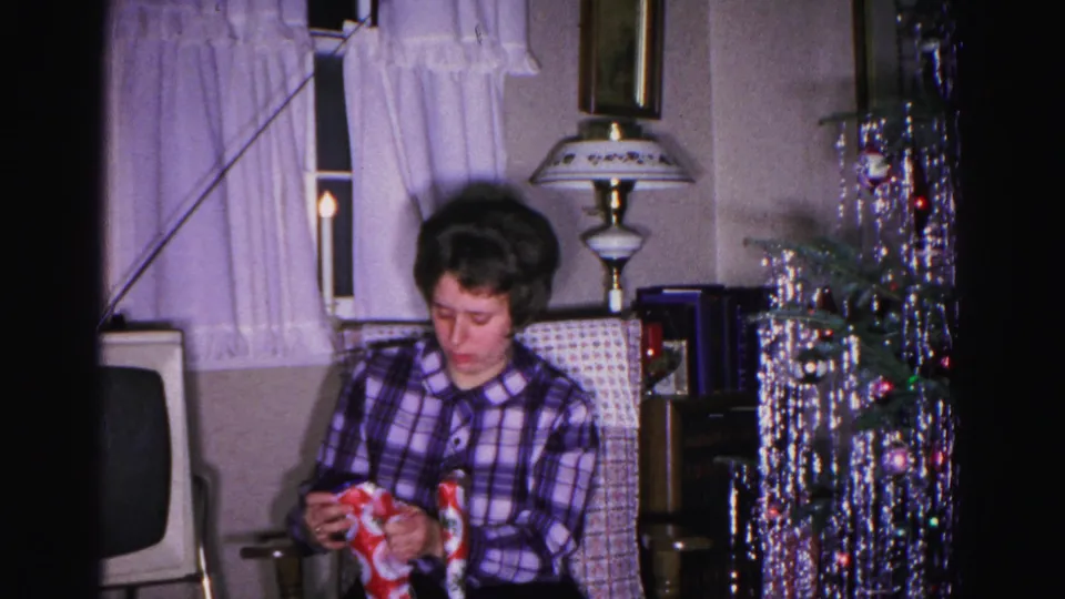 Woman still focused on her gift in a 1960s American living room; aluminum tinsel tree and period CRT television visible in background.