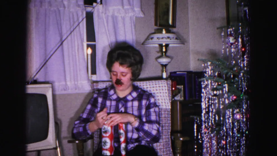 Woman in plaid shirt unwrapping a gift in a 1962 home interior; boxy vintage television at frame left, silver tinsel Christmas tree and table lamp behind her, curtained window above.