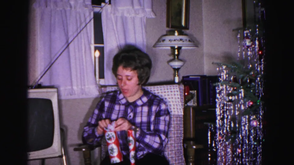 Woman in plaid shirt focused on unwrapping a gift in a 1962 Maryland home; boxy vintage TV, ornate lamp, silver tinsel Christmas tree, and white curtained window visible in background.