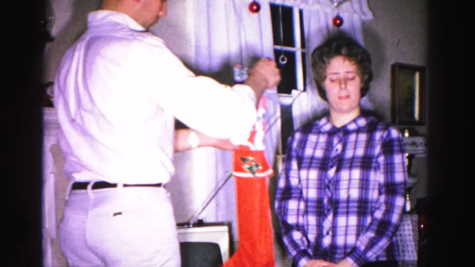 A man in a white shirt and a woman in a plaid blouse hang a red Christmas stocking in a 1962 domestic interior, Hagerstown, Maryland.