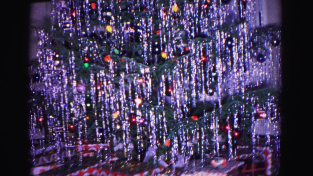 Close-up of a heavily tinseled Christmas tree with colored ball ornaments and small lights in a dim domestic interior.