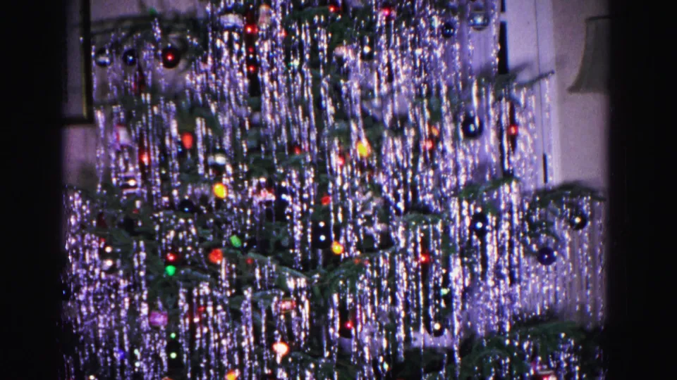 8mm home movie from 1962 showing a Christmas tree with heavy silver tinsel and colored ornaments; white painted wall and interior doorframe in background.