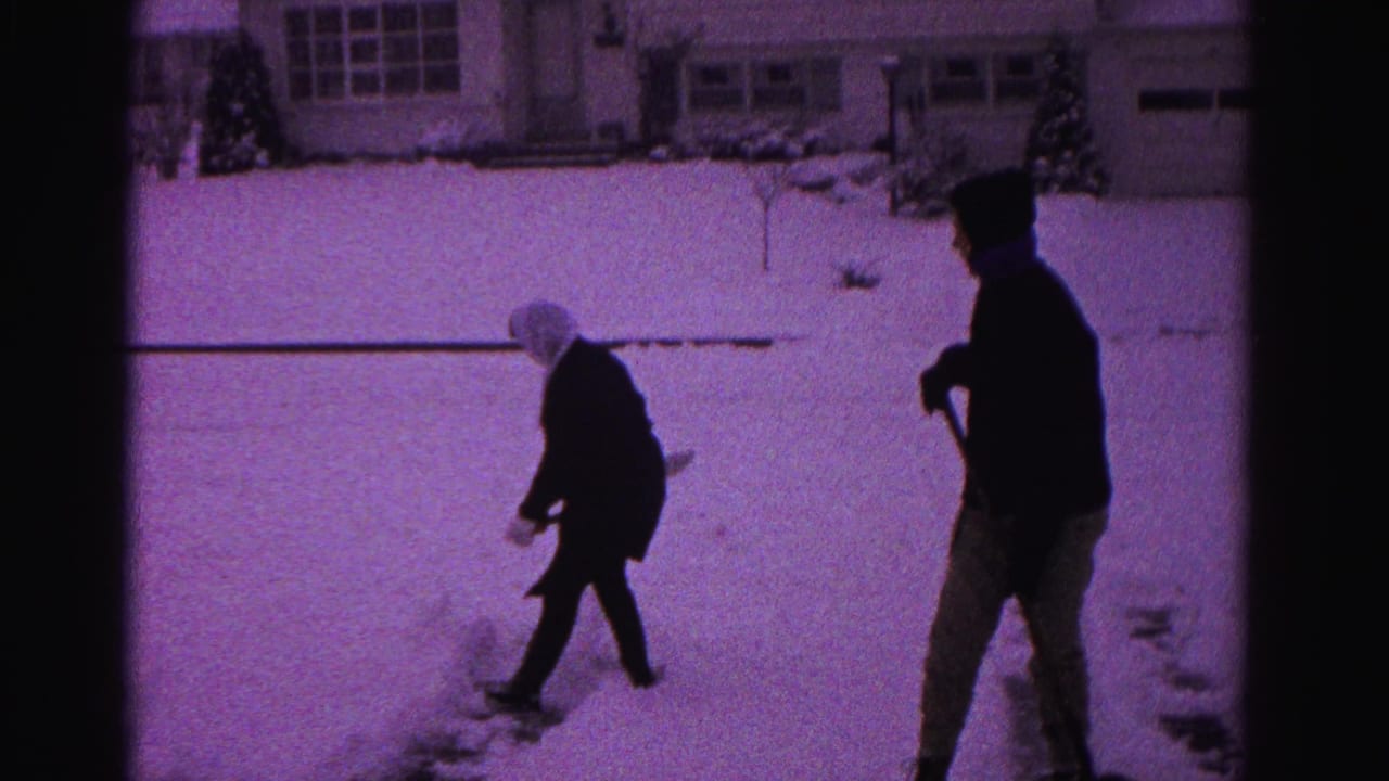 Two adults shoveling snow in a suburban yard with a house visible in the background; strong purple tint from aged 8mm film.