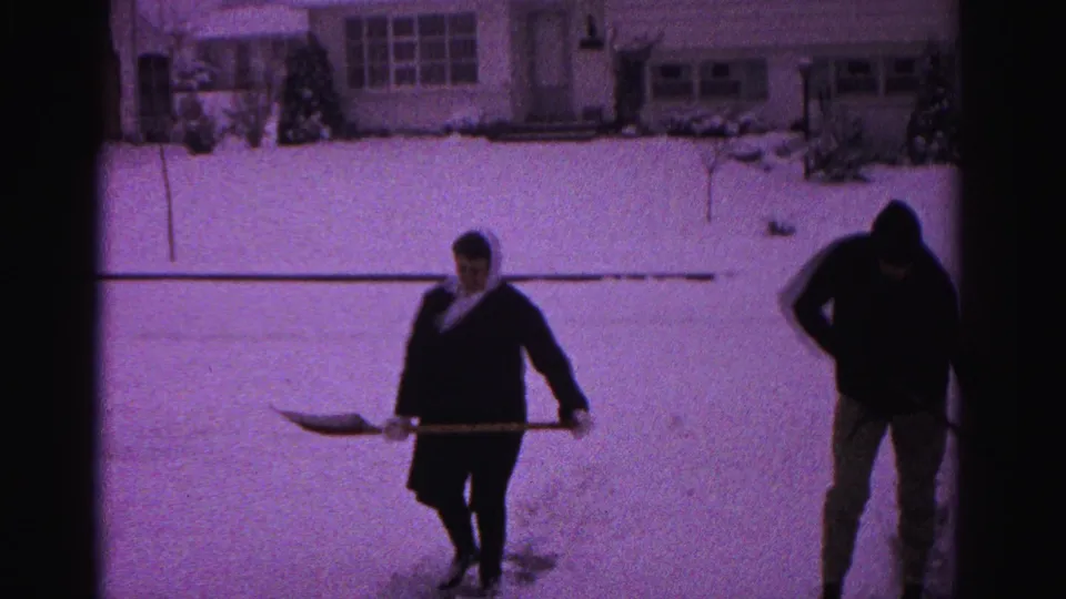 A woman in a head scarf and a man in a hooded jacket pause with shovels facing the camera in a snow-covered suburban yard, circa 1962.