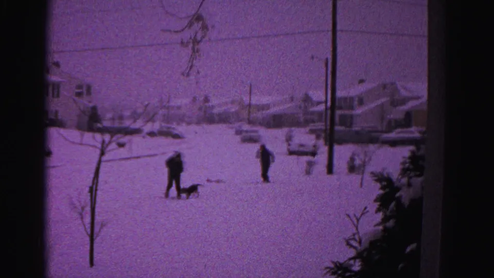 A wide establishing shot of a snow-covered suburban neighborhood, possibly Hagerstown, Maryland circa 1962, with distant figures, a dog, bare trees, and a utility pole.