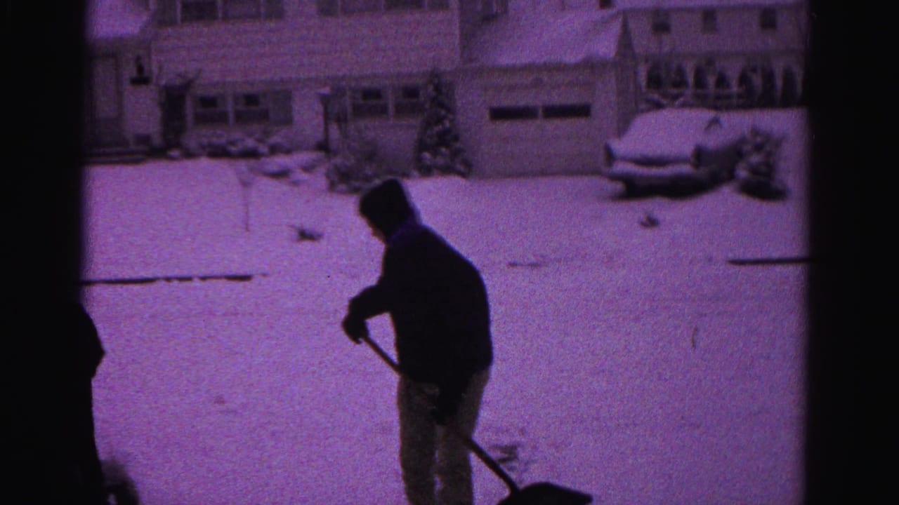 One person in a dark winter coat shoveling snow in a suburban driveway, strong purple color cast from aged 8mm film.