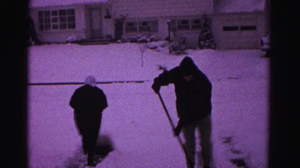 Two individuals clearing snow from a suburban driveway on 8mm film, likely Hagerstown, Maryland, circa 1962; shorter figure appears to wear a light-colored head covering.