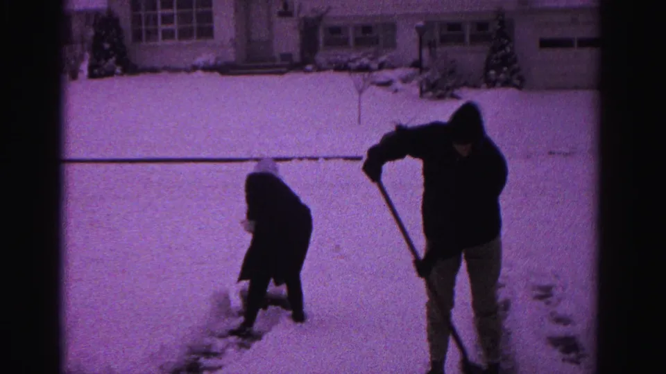 Two people clearing snow from a suburban driveway in this 8mm frame from circa 1962; background shows snow-covered houses and what appear to be evergreen shrubs.