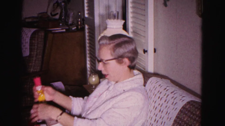 Older woman with glasses and gray hair handles what appears to be a small colored object in a mid-century living room; holiday tinsel visible, circa 1962 Hagerstown, Maryland.
