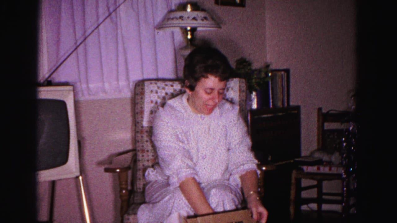 Young woman in white nightgown seated in wicker armchair, looking down at a gift box; vintage TV cabinet and table lamp visible in background.