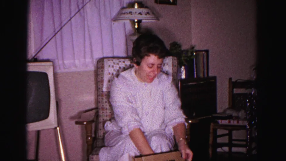 Young woman in white nightgown beginning to open a Christmas gift in a domestic living room, circa 1962 Hagerstown, Maryland; early-sixties television cabinet and ornate lamp behind her.