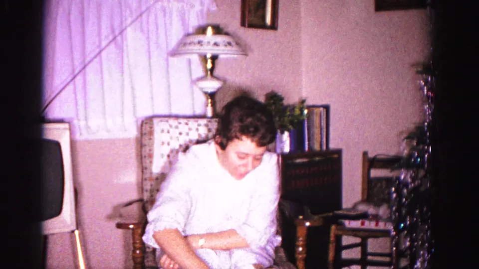 Young woman leaning forward to open a Christmas present in a domestic 1962 interior; decorative lamp, metallic tinsel, and greenery visible behind her.