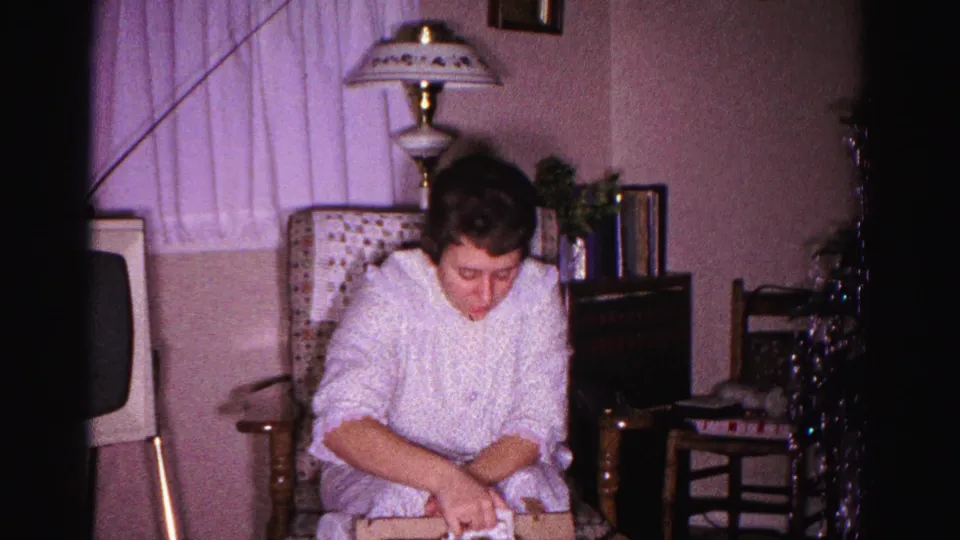 Young woman examining an unwrapped Christmas gift, circa 1962 Hagerstown, Maryland; metallic tinsel decorations and boxy television cabinet visible in background.