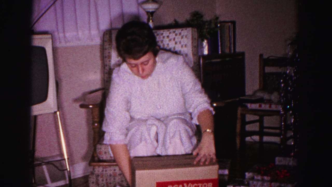 Woman in white robe with hands on a closed RCA Victor gift box in a patterned armchair; 1962 home movie, Hagerstown, Maryland.