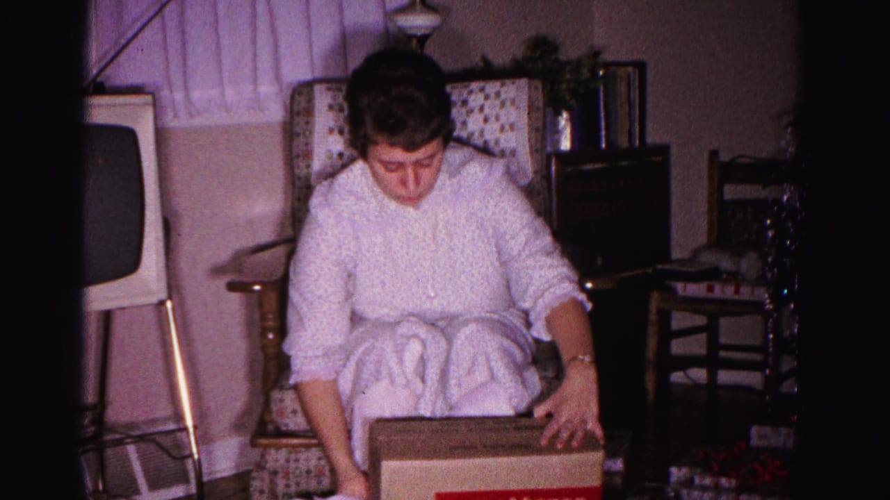 Woman in white robe seated in patterned armchair looking down at RCA Victor box on her lap; vintage television visible at right.