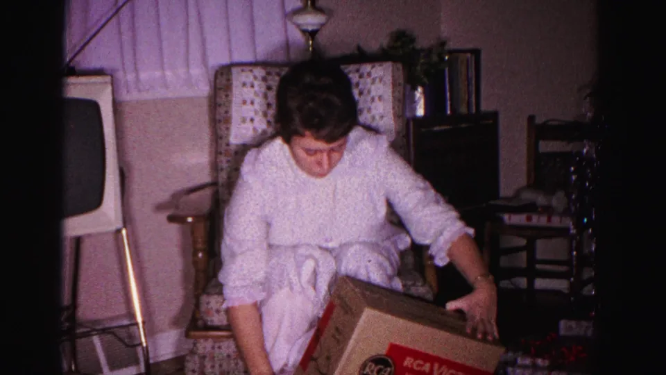 Woman in white robe lifting open an RCA Victor box during a birthday celebration; 8mm home movie, 1962, Hagerstown, Maryland.