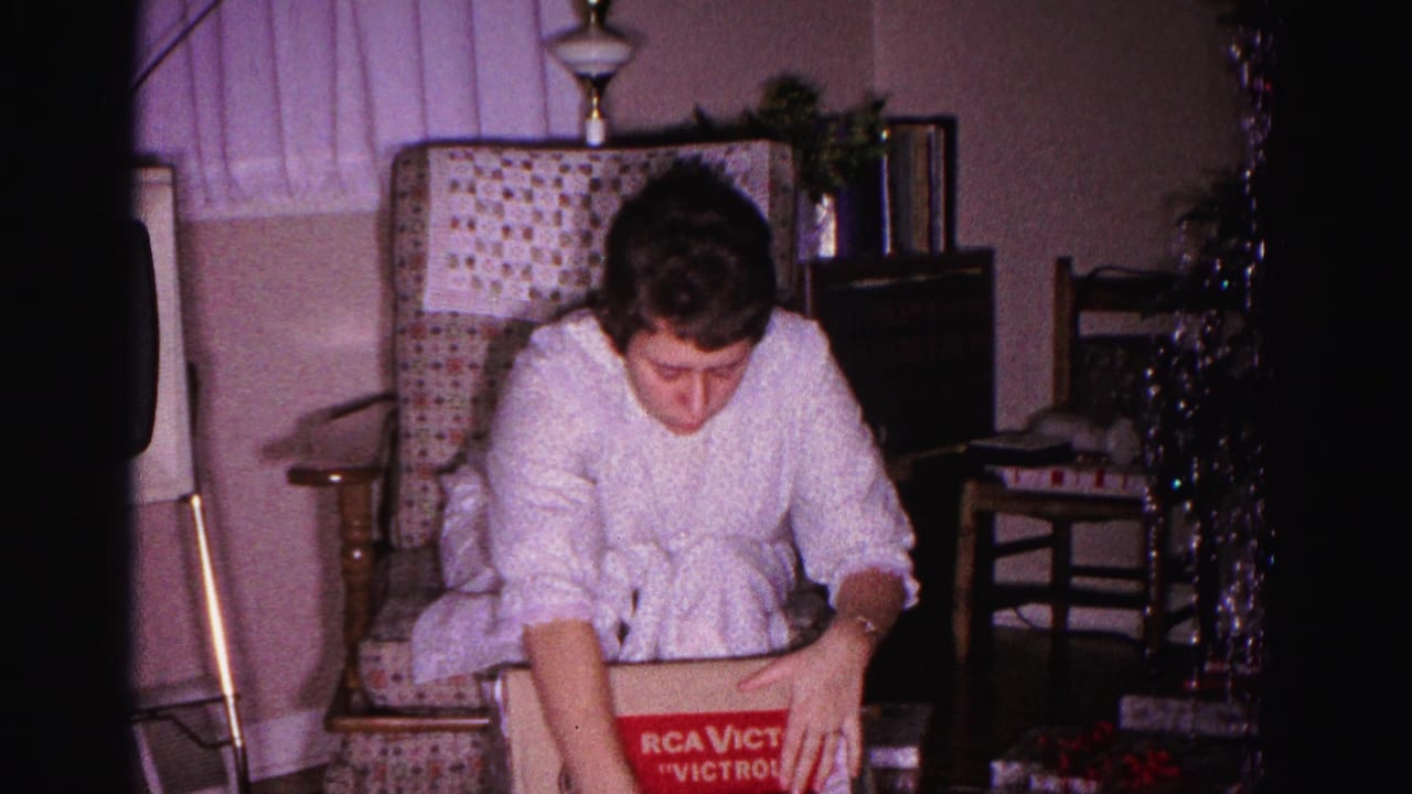 Woman in white housecoat seated in armchair; RCA Victor Victrola box with red-and-white label visible at right, tinsel garland in background.