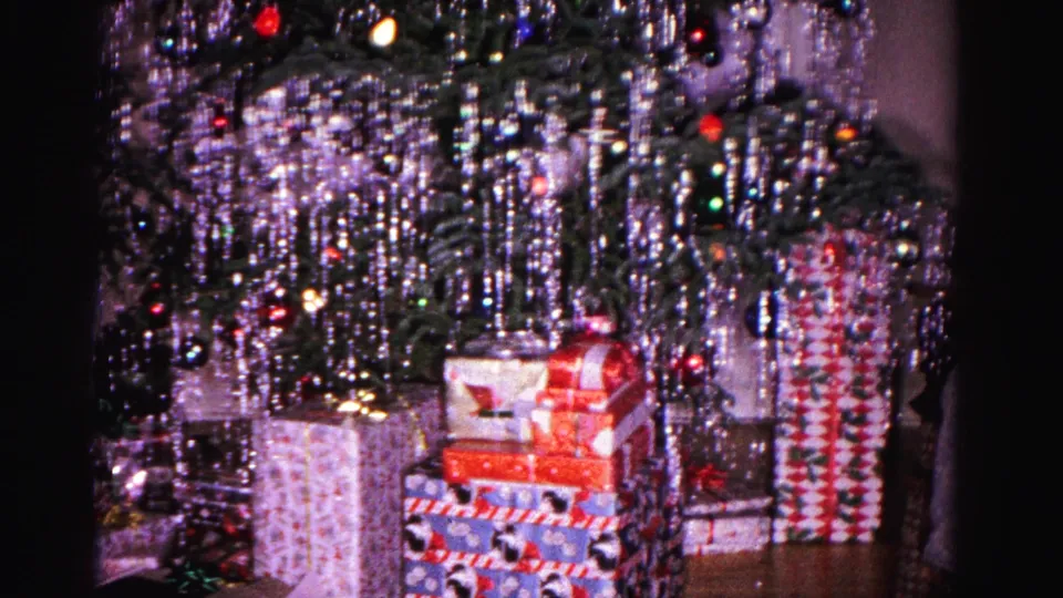 1962 Hagerstown home-movie frame showing the lower branches of a heavily tinsel-covered Christmas tree and multiple wrapped gifts stacked on the floor.