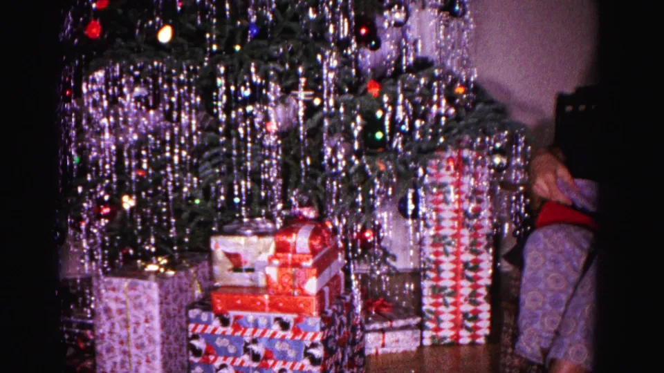 1962 Hagerstown interior frame showing a tinsel-covered Christmas tree, stacked wrapped gifts, and a partially visible seated person at the right edge.