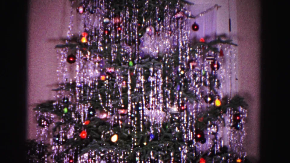 1962 8mm home movie, closer framing of the Christmas tree mid-section showing silver tinsel, multicolored ornaments, and an interior white door in background.