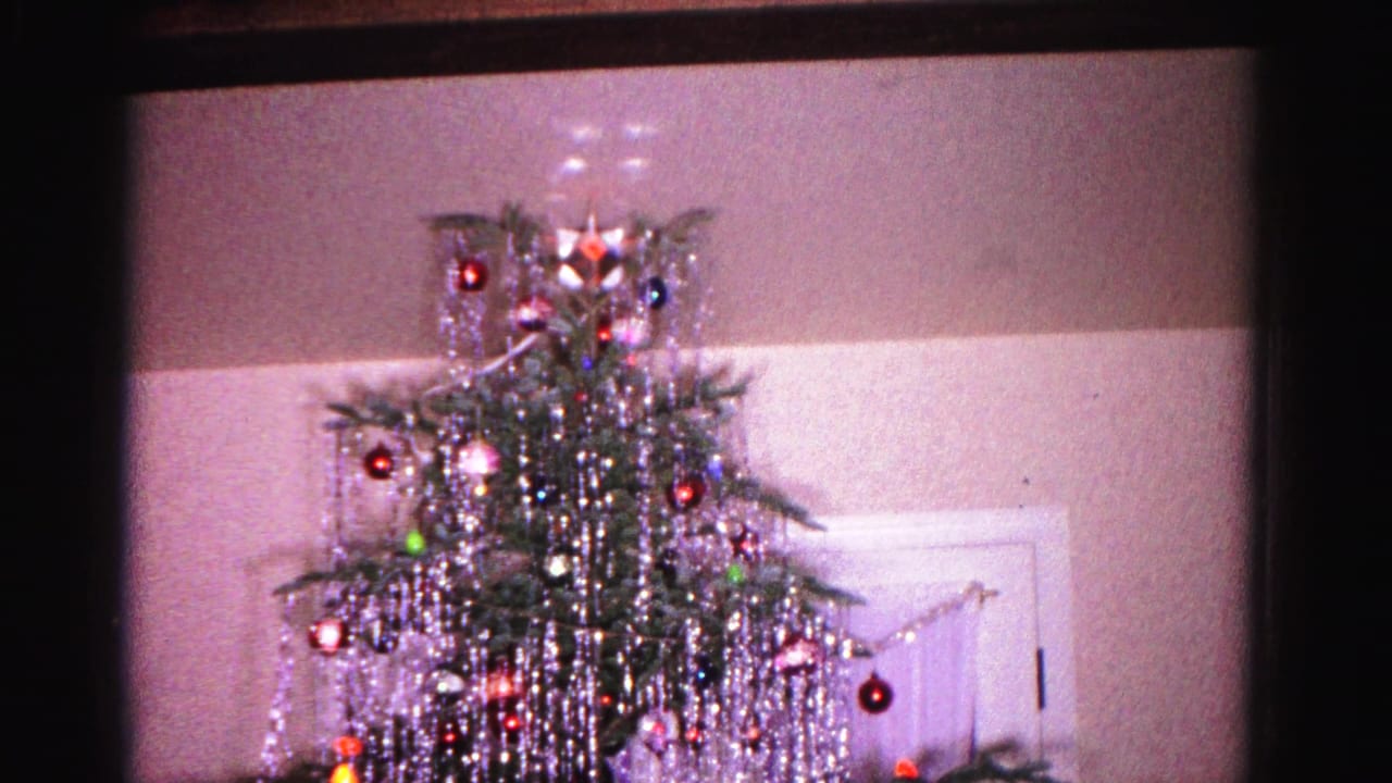 Christmas tree covered in long silver tinsel strands and round ornaments, star topper at apex, white paneled door behind tree, 8mm film frame borders visible.
