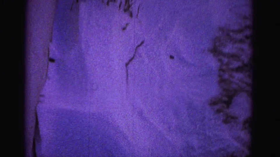 8mm frame circa 1962 showing a deep snow drift pressed against a residential building wall; dry plant stems in the foreground; pronounced purple-violet film color shift.
