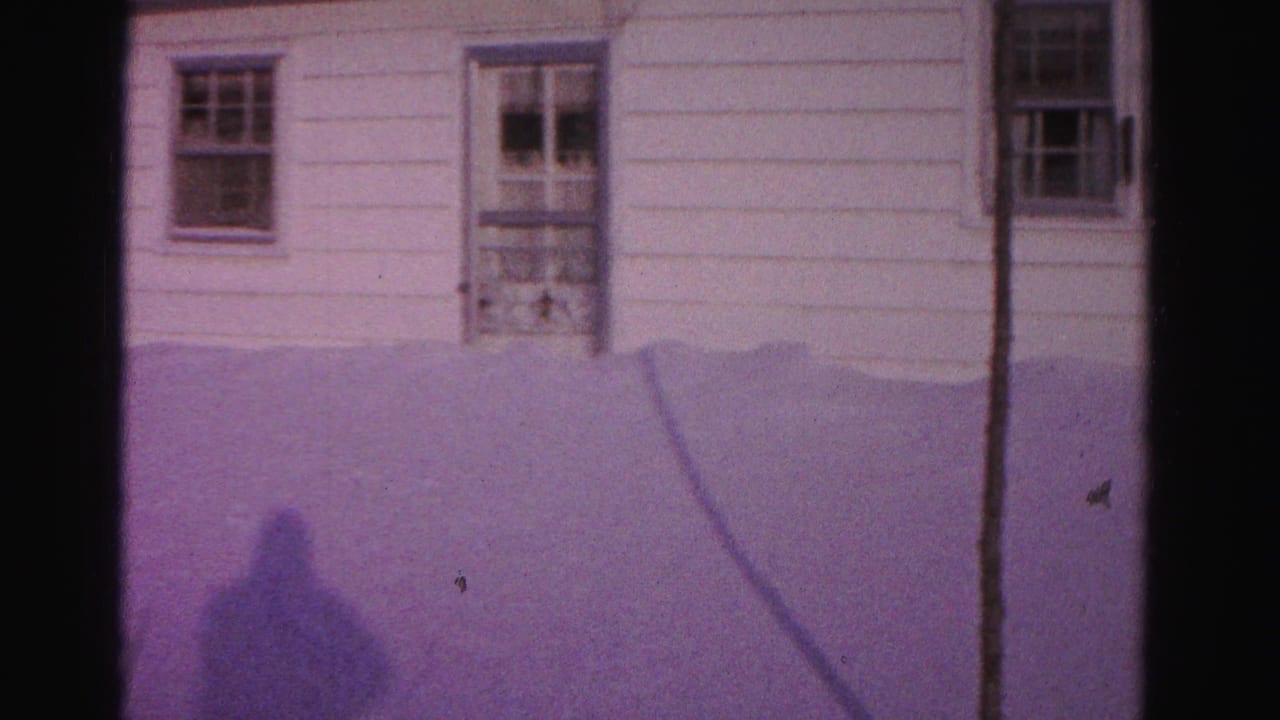 Shadow of a person cast on deep snow in front of a white clapboard house with a screened storm door, filmed on 8mm with a purple tint.