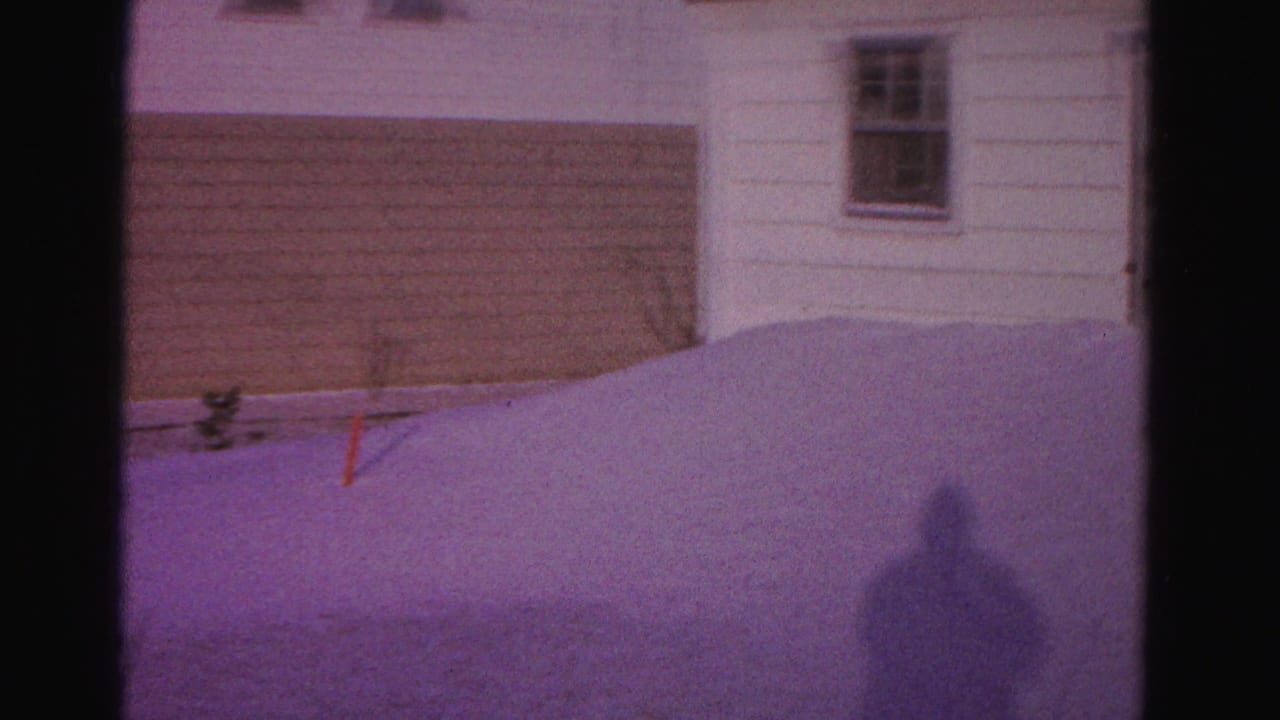 Pan reveals a tan-sided outbuilding beside a white house exterior; filmmaker's shadow on snowdrift and a small red stake in the snow; Hagerstown, Maryland, circa 1962, 8mm.