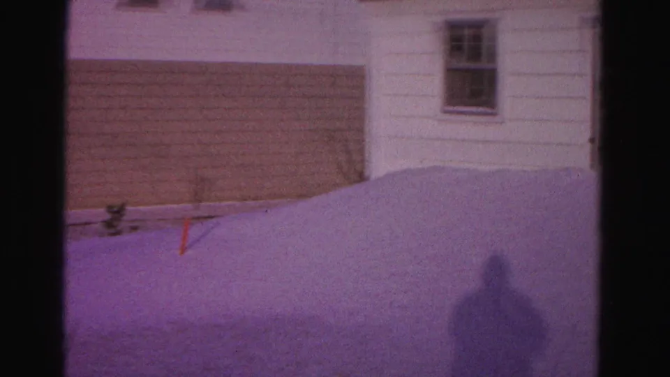 Pan reveals a tan-sided outbuilding beside a white house exterior; filmmaker's shadow on snowdrift and a small red stake in the snow; Hagerstown, Maryland, circa 1962, 8mm.