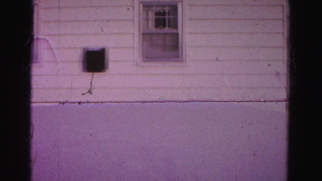 8mm home movie, c.1962, Hagerstown, Maryland — white clapboard house wall with double-hung windows, wall-mounted dark box, and deep snow; purple dye-shift and emulsion scratch visible.