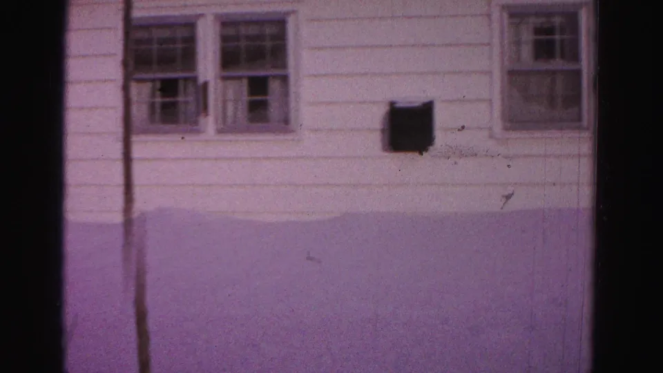 8mm home movie, c.1962, Hagerstown, Maryland — white-sided house wall with three double-hung windows and wall-mounted dark box; deep snow in foreground and aged purple dye-shift throughout.