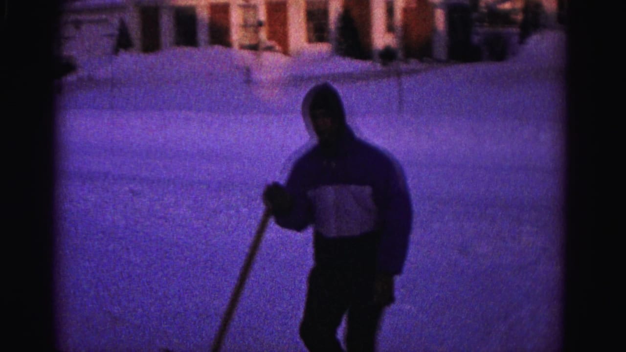 Hooded person holding a snow shovel in close-up, snow-covered surroundings, heavy purple color cast from aged 8mm film.