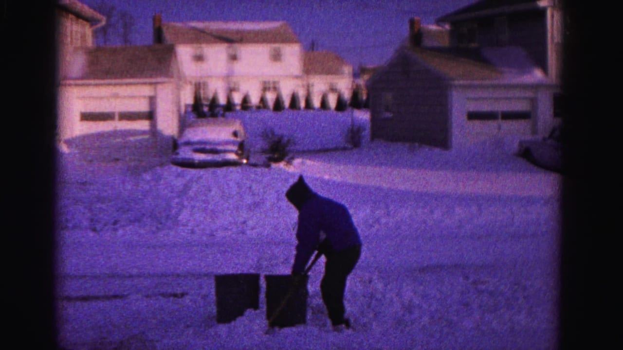 Person in hooded jacket bent forward shoveling snow, with snow-covered houses, detached garage, hedgerow, and partial vintage car visible in background.