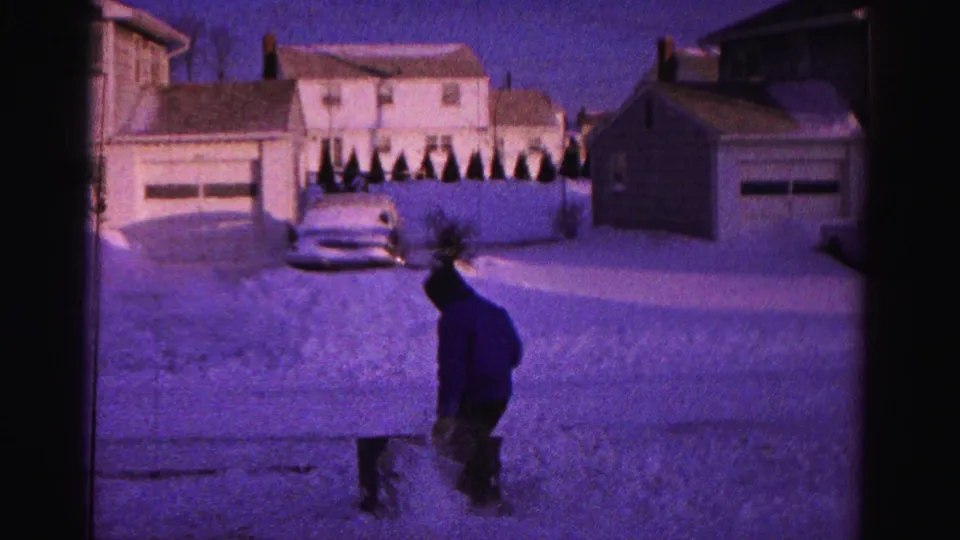A hooded figure stands in a heavily snow-covered residential street in what is noted as Hagerstown, Maryland, circa 1962, with white clapboard houses, detached garages, and a vintage automobile at left on 8mm film.