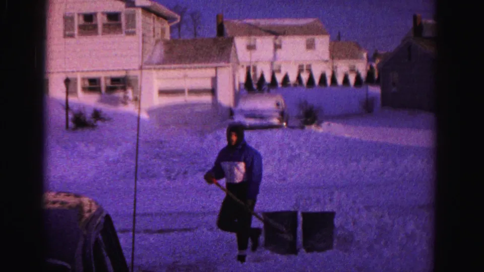 A hooded figure holds a snow shovel beside what appear to be buckets or containers in a snow-covered yard, Hagerstown, Maryland, circa 1962, with suburban houses and garage visible in the background on aged 8mm film.