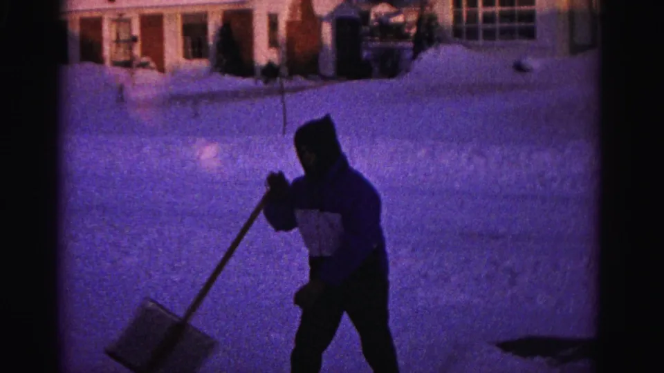 1962 8mm footage of a person in a hooded jacket shoveling snow in what appears to be Hagerstown, Maryland, with suburban houses and deep snow banks behind them.