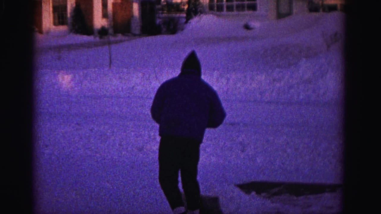 Hooded man standing upright on snow-covered driveway, purple-tinted 8mm footage, residential structures visible in background.