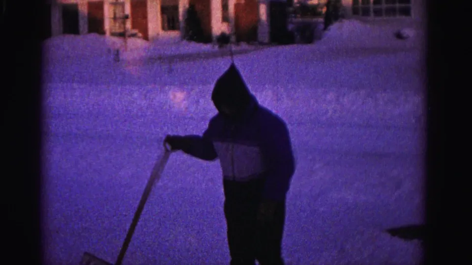Closer shot of hooded man actively shoveling with shovel handle visible, warm house lights contrast with purple snow, 8mm home movie circa 1962.
