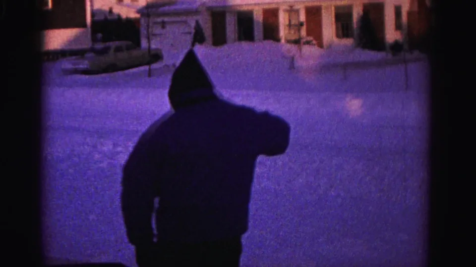 Rear view of hooded man on snow-covered driveway with lit residential houses in background, possible parked vehicle visible, 8mm home movie circa 1962.